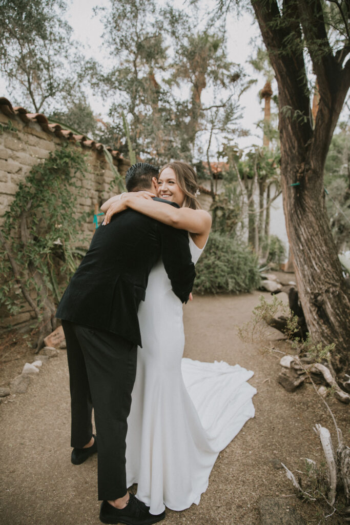There is a bride hugging the groom in a white dress and the groom is in a dark green tux. they are located in a greenery area on a dirt road. The photo is taken behind the groom but cannot see the grooms face but shows the brides face and she is smiling. 